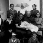 
						The couple holding him are his godmother Mrs Alina Skupinska and his godfather Polish Army Major Dymowski. His mother is sitting at the major's left and at her left is Chaplain Tomasik who baptized him.					