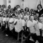 
						Children photographed at mealtime in one of the orphanages in Persia. Counterclockwise standing behind the children - second woman is Mrs Mocarska who later emigrated to the US from England; to her right is my godmother Mrs Alina Skupinska who emigrated to Argentina and from there to Montreal, Canada; fifth person counterclockwise behind the children is my mother Wiktoria Morawska.					